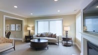 This two-story residence combines timeless design with a warm, understated aesthetic throughout its living spaces. The living room showcases a carefully curated interior, highlighted by smooth ceilings with recessed lighting and elegant crown molding that frames the room with architectural detail. Neutral, warm-toned walls create a calming backdrop that pairs naturally with the plush, patterned carpeting underfoot.

A central tufted sofa anchors the seating area, flanked symmetrically by dark-stained round side tables, each topped with classic table lamps that contribute soft, ambient lighting. A substantial dark wood oval coffee table serves as a focal point, lending depth and contrast to the lighter elements throughout the room. A wide window with white horizontal blinds allows natural light to filter in while maintaining privacy, enhancing the room's airy and open feel.

The thoughtful balance of dark wood furniture against light walls and soft carpeting gives the space a polished, cohesive character. The overall ambiance is one of quiet sophistication — designed to feel both elegant and comfortable across both floors of the home.