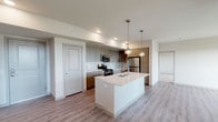 Located at Townsite Promenade in Camarillo, California, this modern single-floor residence offers a thoughtfully designed layout with contemporary finishes throughout. The open-concept kitchen serves as the heart of the home, anchored by a sleek white quartz island with an integrated sink and pendant lighting overhead. Two-tone cabinetry in warm wood and gray tones lines the walls, complemented by a subway tile backsplash, stainless steel appliances including a refrigerator, gas range, and built-in microwave. Light wood-style flooring flows through the kitchen and living areas, creating a cohesive and clean aesthetic. The living space is bright and open, benefiting from multiple windows that invite natural light and offer views of the surrounding Camarillo community. Bedrooms feature plush carpet flooring and generously sized windows that continue to bring in natural light. The bathroom includes a full soaking tub and shower combination with crisp white tile surround, a quartz-topped vanity, and modern chrome fixtures. An additional bathroom provides functional convenience. The overall design palette of neutral whites and warm wood tones lends the space a fresh, contemporary ambiance suited to modern living.