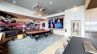This contemporary amenity lounge is thoughtfully designed to serve as a dynamic social and entertainment hub. The space is anchored by a sleek black pool table with a bold orange felt surface, positioned beneath a sculptural starburst chandelier that serves as a striking focal point overhead. Recessed lighting complements the statement fixture, ensuring the space is well-illuminated throughout.

A large-format flat-screen display mounted on a crisp white wall provides an immersive viewing experience, flanked by built-in cabinetry that offers both storage and a clean, streamlined aesthetic. Pool cues are neatly racked nearby for easy access.

A dramatic mountain landscape mural with vivid red neon accents commands an entire wall, adding energy and visual depth to the lounge. A casual seating area with plush cushions and accent ottomans is nestled beneath the mural, creating an inviting relaxation zone. A long conference-style table with modern chairs along one side provides a functional workspace or gathering area. Neutral-toned carpet and light tile flooring tie the design together, contributing to a polished, modern ambiance suited for both relaxation and social interaction.