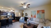 This single-floor space offers an open-concept layout that brings together the living, dining, and kitchen areas in a cohesive and functional design. The living room serves as the heart of the space, featuring a ceiling fan with integrated lighting that complements the bright, neutral walls. Large abstract artwork in warm tones of orange, teal, and white adds a vibrant focal point to the room, while plush carpeting underfoot creates a comfortable and inviting atmosphere. The living area is furnished with a spacious sectional sofa and coordinating armchair, centered around a rich, dark coffee table that anchors the space. A stylish arc floor lamp with a round mirror accent piece adds an artistic touch. Adjacent to the living room, the dining area accommodates a full dining table and chairs, with hardwood-style flooring that distinguishes it from the carpeted living space. The kitchen is visible beyond, featuring dark cabinetry and black appliances, offering a classic and practical cooking environment. The open sightlines throughout the space create a sense of generous square footage, making it feel airy and well-connected. The overall ambiance is warm, welcoming, and thoughtfully designed for everyday living and entertaining.