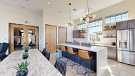 This single-floor space is thoughtfully designed in a contemporary style, combining an open-plan kitchen, dining area, and lounge into one cohesive, light-filled environment. The soaring ceilings amplify the sense of openness, while clerestory windows and large sliding glass doors flood the interior with abundant natural light and offer direct views of an outdoor patio area.

The kitchen is a standout feature, showcasing a striking combination of navy blue lower cabinetry and dark wood upper cabinets, accented by a crisp white quartz countertop. A large central island with bar seating for several occupants serves as a social hub, anchored beneath two sculptural brass and globe pendant lights. Open wooden shelving, a white subway tile backsplash, a gold-finished faucet, and built-in appliances complete the well-appointed kitchen.

The dining area includes a marble-topped table beneath a warm gold pendant light, complemented by geometric wall art and direct exterior access. The lounge area features a large sliding glass wall opening to a spacious patio, navy built-in cabinetry with gold hardware, and a mounted display screen. Light tile flooring and patterned area rugs unify the entire space, creating a refined, welcoming atmosphere.