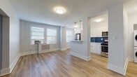 This thoughtfully renovated single-floor unit offers a clean, modern aesthetic throughout. The open-concept layout seamlessly connects the living area to the kitchen, creating a spacious and airy feel. Light hardwood flooring flows continuously across the main living space, complementing the soft gray walls and bright white trim.

The living area is well-lit by multiple windows fitted with classic horizontal blinds, allowing ample natural light to fill the room. A radiator cover and window air conditioning unit reflect the unit's practical character. A striking navy blue accent wall anchors one corner of the living space, adding a bold design element.

The kitchen is a standout feature, defined by crisp white shaker-style cabinetry, a bold navy herringbone tile backsplash, and a suite of stainless steel appliances including a refrigerator, gas range, microwave, and dishwasher. A peninsula with pendant lighting above creates a natural divide between the kitchen and living area while offering additional counter space.

The bathroom is elegantly finished with deep navy subway tile extending from floor to ceiling in the shower and tub surround, paired with slate gray floor tiles. A window above the tub provides natural light, while clean white fixtures and modern hardware complete the look. The overall design of the unit balances contemporary style with practical, well-chosen finishes throughout.