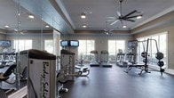 This fitness center is designed to provide a comprehensive workout experience in a bright, well-appointed setting. The space is laid out on a single floor and features a thoughtful arrangement of both cardio and strength training equipment. Prominently displayed along one side are multiple treadmills and elliptical machines, positioned to take full advantage of the large windows that line the walls, allowing ample natural light to fill the room and offering pleasant outdoor views during workouts. Body-Solid strength training machines occupy the opposite side of the space, complemented by full-length mirrored walls that enhance the sense of openness and allow users to monitor their form. The flooring consists of durable, dark rubberized material well-suited for a high-activity environment. Overhead, a tray ceiling with crown molding adds an architectural refinement, while two large multi-blade ceiling fans ensure consistent air circulation throughout the room. Recessed lighting supplements the natural light, keeping the space well-illuminated at all times. Multiple wall-mounted flat-screen televisions are positioned for easy viewing from the cardio equipment, adding an entertainment element to the workout experience. The overall ambiance is clean, spacious, and motivating.