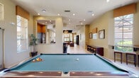 This single-floor community clubhouse is thoughtfully designed to offer a variety of social and recreational amenities within a cohesive, open layout. The space is finished throughout with large-format light tile flooring that enhances its bright, airy atmosphere.

The game room serves as a central highlight, featuring a full-size pool table set atop a carpeted area, flanked by dual flat-screen televisions mounted beneath decorative hood-style surrounds. Bar-height stools line the perimeter, providing casual seating, while tall windows flood the area with natural light and views of the surrounding greenery.

Adjacent to the game room is a well-equipped kitchen with dark cabinetry, granite countertops, stainless steel built-in ovens, and a large island with bar seating beneath striking pendant lights. The kitchen opens directly into the main gathering area, creating a seamless flow ideal for entertaining.

The lounge area features a comfortable seating arrangement with a sofa and armchairs centered around an entertainment center, beneath a distinctive dark wood tray ceiling with a ceiling fan. Tall decorative windows frame the space and invite abundant natural light.

A windowed counter nook with granite surfaces and bar stools offers an intimate spot for conversation or casual dining, overlooking the outdoor landscape. The overall ambiance is warm, welcoming, and well-suited for community gatherings.