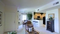 This two-floor residence is designed with an open and connected layout that promotes a comfortable, functional living environment. The main living area is carpeted and features crown molding along the ceiling, adding a classic architectural detail to the space. Track lighting runs overhead, providing flexible illumination throughout the room.

The living and dining areas flow seamlessly into one another, with a plaid upholstered armchair and a dining table with wooden chairs creating distinct yet connected zones. Lush indoor plants add greenery and warmth to the space. A bright window at the far end of the dining area allows natural light to fill the room.

The kitchen is positioned adjacent to the main living space and features light wood cabinetry that extends to the upper walls, dark granite countertops, a black refrigerator, and a built-in microwave. A raised bar counter with bar stools provides additional seating and serves as a natural boundary between the kitchen and dining areas. The kitchen is finished with tile flooring, contrasting the carpet of the living spaces. Overall, the residence offers a bright, welcoming ambiance with thoughtful design throughout.