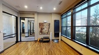This single-floor fitness facility is thoughtfully designed to support a wide range of workout routines in a bright, welcoming environment. The space is anchored by a generously sized open exercise area finished with warm, striped bamboo flooring that lends a natural, energizing feel underfoot. Large, floor-to-ceiling windows line the exterior walls, flooding the interior with abundant natural light and offering views of the surrounding greenery and streetscape outside.

The main studio area is well-equipped for functional and group training, featuring medicine ball racks, stability domes, resistance bands, yoga mats, and low workout benches. A large wall-mounted flat-screen display serves as a focal point, ideal for guided workout sessions, while a ceiling fan overhead helps maintain comfortable airflow throughout. Decorative wall art adds a thoughtful aesthetic touch to the space.

Glass-paneled doors connect the studio to an adjacent weight room, visible through the dividers and housing a range of strength training machines and equipment. Recessed lighting throughout ensures the space is evenly and efficiently illuminated regardless of the time of day. The overall design balances functionality with a clean, modern aesthetic, creating an inviting atmosphere suitable for yoga, stretching, cardio, and strength training alike.