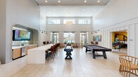 This contemporary community amenity space makes a striking first impression with its soaring double-height lobby, anchored by a sculptural black spiral staircase featuring an intricate geometric railing design. A gold sunburst chandelier crowns the space above a sleek reception desk with wood-panel accents, while large floor-to-ceiling windows and glass doors flood the area with natural light. Light-toned tile flooring flows throughout the ground level, lending the interiors a clean, open feel.

Adjacent to the entry, a well-appointed lounge area features a curated mix of seating, a coffee bar with light wood cabinetry, and a dedicated workspace zone with semi-private booth seating. Tall stone-clad accent walls and modern pendant lighting add warmth and texture throughout. A separate lounge with expansive windows and outdoor access offers a comfortable bar-height counter, statement wall art, and panoramic views of lush greenery.

The upper mezzanine level—accessible via the spiral staircase—provides a relaxed retreat with patterned carpeting, hanging egg chairs, bean bag seating, and a bookshelf nook. Additional amenities include a fully stocked convenience market with refrigerated beverages and pantry items, a serene salon-style waiting area with hardwood flooring and blue upholstered chairs, and a dedicated package locker room for secure parcel delivery.