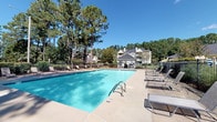 Located at 215 Valencia Dr in Jacksonville, North Carolina, this outdoor community amenity space offers residents a well-appointed retreat designed for relaxation and social gatherings. The centerpiece of the space is a generously sized swimming pool with crystal-clear blue water, surrounded by a wide concrete pool deck lined with multiple lounge chairs — ideal for sunbathing on bright, clear days. Tall pine trees frame the perimeter, providing natural shade and a sense of seclusion while maintaining an open, airy atmosphere under the North Carolina sky. Access to the pool area is provided through a distinctive white lattice-style gate, which complements the architectural character of the surrounding community buildings. Adjacent to the pool, a white-painted open-air gazebo serves as a covered gathering space. The gazebo features a warm wood-beam vaulted ceiling, decorative cross-lattice railings, a gas grill, and a picnic table — making it a functional outdoor entertaining area. A fire extinguisher is mounted within the gazebo, reflecting attention to safety. The surrounding landscaping includes manicured hedges, ornamental grasses, and mature trees that contribute to the green, park-like setting of this community outdoor space.