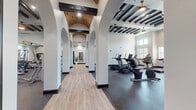 This expansive fitness center combines striking architectural design with a comprehensive array of exercise equipment. The space is defined by a dramatic arched colonnade corridor with a vaulted brick ceiling and bold dark beams, creating an inviting passage between workout zones. Light hardwood flooring in the corridor transitions to durable rubber flooring throughout the equipment areas, providing both aesthetic contrast and functional support.

The cardio zone is well-equipped with True treadmills, StairMaster steppers, HIIT rowing machines, and stationary bikes arranged to maximize space and comfort. Large windows along the walls flood the area with natural light and offer views of the surrounding outdoor landscape. Elevated clerestory windows further enhance the sense of openness and airiness.

The strength training area features a robust selection of equipment including cable machines, power racks, adjustable benches, a full dumbbell rack, and stability balls. Full-length mirrors are strategically placed to assist with form and to amplify the sense of space. Exposed dark wood beam ceilings with recessed and pendant lighting add warmth and architectural character throughout.

The overall ambiance is one of refined functionality — a fitness facility that balances upscale design with practical, well-organized workout spaces suited for a wide range of fitness routines.
