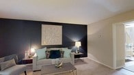 This two-floor space makes a strong design statement through its thoughtfully curated interiors. The living area serves as a focal point, anchasing a bold, dark accent wall that creates a striking contrast against the neutral-toned walls and light ceiling. A large, geometric wooden wall art piece commands attention above the sofa, adding texture and visual interest to the space. Warm ambient lighting from coordinating table lamps on either side of the seating area casts a cozy, inviting glow throughout the room. The living area is furnished with a comfortable, neutral-toned sofa adorned with teal and dark accent pillows, complementing the overall color palette of teal, black, and gold. A sleek, dark coffee table anchors the seating arrangement, while plush carpeting underfoot adds warmth and comfort. The carefully layered lighting design enhances the sophisticated ambiance of the space. The two-floor layout offers additional living areas beyond what is visible, providing ample room for varied functional spaces. Overall, the interior design reflects a contemporary aesthetic with a deliberate use of contrast, texture, and accent tones to create a polished and inviting environment.