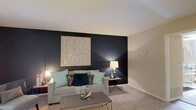 This two-floor space makes a strong design statement through its thoughtfully curated interiors. The living area serves as a focal point, anchasing a bold, dark accent wall that creates a striking contrast against the neutral-toned walls and light ceiling. A large, geometric wooden wall art piece commands attention above the sofa, adding texture and visual interest to the space. Warm ambient lighting from coordinating table lamps on either side of the seating area casts a cozy, inviting glow throughout the room. The living area is furnished with a comfortable, neutral-toned sofa adorned with teal and dark accent pillows, complementing the overall color palette of teal, black, and gold. A sleek, dark coffee table anchors the seating arrangement, while plush carpeting underfoot adds warmth and comfort. The carefully layered lighting design enhances the sophisticated ambiance of the space. The two-floor layout offers additional living areas beyond what is visible, providing ample room for varied functional spaces. Overall, the interior design reflects a contemporary aesthetic with a deliberate use of contrast, texture, and accent tones to create a polished and inviting environment.