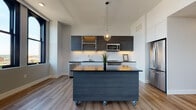 Located on Church Street in the heart of downtown Akron, Ohio, this single-floor unit showcases a thoughtfully designed contemporary interior with high-end finishes and an open, airy layout. Light wood-tone flooring runs throughout, tying each space together cohesively.

The kitchen is a standout feature, outfitted with dark wood-grain upper cabinetry paired with clean white lower cabinets and black countertops. Under-cabinet lighting, a white subway tile backsplash, and stainless steel appliances — including a dishwasher, range, and built-in microwave — complete the culinary space. A large moveable kitchen island on casters adds versatile prep and entertaining space, topped with a matching dark surface and highlighted by a sleek globe pendant light above.

The living area is generously proportioned, featuring smooth walls, recessed lighting, a ceiling fan, and distinctive arched windows that frame views of the Akron skyline and fill the room with natural light.

The bathroom maintains the modern aesthetic with large-format tile flooring, a wall-mounted rectangular sink, a backlit vanity mirror, a tall built-in storage cabinet, and clean white walls. The overall ambiance of the unit is polished, minimalist, and move-in ready.