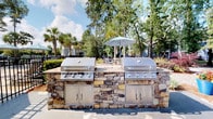 Located at 8439 Dorchester Rd in North Charleston, South Carolina, this outdoor amenity space offers a well-appointed communal grilling station designed for gathering and entertaining. The centerpiece of the area is a substantial stone-clad outdoor kitchen structure, housing two large stainless steel built-in gas grills with ample burners and integrated storage cabinets below — ideal for hosting cookouts and community events.

The natural stone surround gives the space a sturdy, refined aesthetic that complements the lush, verdant surroundings. Tall pine and palm trees frame the area, providing a canopy of greenery that enhances the relaxed, outdoor atmosphere characteristic of the South Carolina Lowcountry. Nearby, a shaded seating area with a patio umbrella and chairs offers a comfortable spot for dining and socializing al fresco.

The space benefits from abundant natural light and open-air design, creating an inviting environment throughout the day. The surrounding landscape, featuring manicured hedges and mature trees, adds to the sense of privacy and seclusion. This thoughtfully designed outdoor amenity reflects a commitment to community living, providing residents with a functional and attractive space to enjoy the warm Carolina climate year-round.