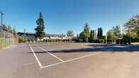 This outdoor recreational space features a well-maintained tennis court with clearly marked playing lines and a regulation net at its center. The court surface is a classic dark asphalt finish, offering a firm and even playing area suitable for year-round use. The facility is fully enclosed by chain-link fencing, which lines the perimeter to keep play contained and provide a defined boundary for the space.

The surroundings are richly landscaped, with tall evergreen trees, columnar conifers, and neatly trimmed hedgerows framing the court on multiple sides. These plantings provide a natural windbreak and visual privacy, contributing to a pleasant and focused playing environment. Court-side lighting posts are visible along the perimeter, suggesting the facility supports play during low-light hours.

Beyond the fencing, a gently sloping grassy lawn leads up to a multi-story residential building featuring balconies and large windows. Additional mature trees, including a striking weeping willow and a towering fir, add character and shade to the broader grounds. The open exposure to the sky ensures ample natural light throughout the day, and the clear, unobstructed overhead views enhance the sense of openness. Overall, this space offers a well-appointed and inviting setting for recreational tennis within a communal residential environment.
