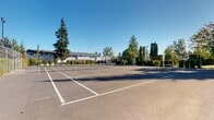 This outdoor recreational space features a well-maintained tennis court with clearly marked playing lines and a regulation net at its center. The court surface is a classic dark asphalt finish, offering a firm and even playing area suitable for year-round use. The facility is fully enclosed by chain-link fencing, which lines the perimeter to keep play contained and provide a defined boundary for the space.

The surroundings are richly landscaped, with tall evergreen trees, columnar conifers, and neatly trimmed hedgerows framing the court on multiple sides. These plantings provide a natural windbreak and visual privacy, contributing to a pleasant and focused playing environment. Court-side lighting posts are visible along the perimeter, suggesting the facility supports play during low-light hours.

Beyond the fencing, a gently sloping grassy lawn leads up to a multi-story residential building featuring balconies and large windows. Additional mature trees, including a striking weeping willow and a towering fir, add character and shade to the broader grounds. The open exposure to the sky ensures ample natural light throughout the day, and the clear, unobstructed overhead views enhance the sense of openness. Overall, this space offers a well-appointed and inviting setting for recreational tennis within a communal residential environment.