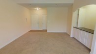 This two-floor property offers a straightforward and functional layout designed to maximize usable living space. The main living area, pictured here, features wall-to-wall beige carpeting that adds warmth underfoot, complemented by smooth, neutral-toned walls that create a bright and inviting atmosphere. Overhead, a flush-mount ceiling light provides consistent illumination throughout the room.

The entryway area is defined by two white paneled doors side by side — one serving as the main entry and the other likely providing access to a closet — framed against a backdrop of hardwood or hard-surface flooring that transitions into the carpeted living space. This subtle shift in flooring helps distinguish the entry zone from the main living area. The open layout lends itself to flexible furniture arrangements and a sense of spaciousness.

White baseboards trim the perimeter of the room, adding a crisp finishing detail against the neutral walls. To one side, a partial view suggests additional rooms or spaces that connect to the main living area. The overall ambiance is clean, neutral, and move-in ready, offering a blank canvas for personalization across both floors.