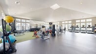 Located at 10801 Heather Ridge Cir in Orlando, FL, this generously sized fitness center offers a comprehensive, single-floor workout facility designed to accommodate a wide range of exercise needs. The space is defined by its striking vaulted ceilings accented by a large skylight, which floods the interior with natural light and creates an open, airy atmosphere throughout.

The facility is thoughtfully organized into distinct workout zones. A dedicated cardio area lines one side of the space with a row of treadmills equipped with individual screens, alongside elliptical trainers and stationary cycling bikes, all positioned near banks of large multi-pane windows that offer views of the surrounding outdoor environment. The two-tone wall design — neutral beige above and a deeper slate blue below — gives the space a clean, polished aesthetic.

The strength and functional training zone features a power rack with a bench press station, a cable crossover machine, a full dumbbell rack backed by floor-to-ceiling mirrors, and a variety of selectorized resistance machines. Green turf flooring demarcates this area, contrasting with the durable rubberized dark flooring throughout the main space. Medicine balls and resistance accessories round out the functional fitness offerings. Wall-mounted televisions are positioned for visibility across the floor, and a vending machine provides convenient on-site refreshments. The overall layout is spacious, well-lit, and designed to support both individual and group fitness activity.