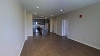 This single-floor space showcases an open-concept design that seamlessly connects the living area to the kitchen, creating a spacious and functional layout. The dark hardwood-style flooring runs throughout, providing a warm, cohesive foundation that contrasts attractively with the neutral beige and gray tones of the walls.

The kitchen is a standout feature, equipped with dark gray cabinetry, stainless steel appliances including a range, microwave, and refrigerator, and a subway tile backsplash that adds texture and visual interest. A central island offers additional workspace and casual seating potential, making it a practical hub for cooking and entertaining.

Recessed lighting is installed throughout the ceiling, providing even, consistent illumination across the open living and kitchen areas. The clean white baseboards and door trim offer a crisp contrast to the darker flooring. Multiple doorways visible toward the rear of the space suggest access to additional rooms, contributing to the overall functionality of the layout. The open floor plan and unobstructed sightlines lend the space a generous, airy feel.