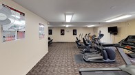 This fitness center offers a comprehensive workout environment spread across a single level. The space is divided into distinct functional zones, each outfitted with a variety of exercise equipment to accommodate different training needs.

The main cardio area features a row of treadmills and elliptical trainers from brands including Matrix, providing ample options for aerobic exercise. Motivational athletic posters and instructional fitness charts are displayed along the neutral-toned walls, adding visual interest while serving as practical references. A wall-mounted television is positioned for easy viewing during workouts.

A separate strength training area houses a multi-station cable machine along with additional resistance equipment, giving users access to a range of resistance-based exercises. Protective rubber mats are placed beneath the equipment throughout the space.

A small seating nook with a bistro-style table and bar stools offers a rest area, accompanied by a wall-mounted air conditioning unit for climate control. Vertical blinds cover the windows, allowing natural light while maintaining privacy. Overhead fluorescent panels provide consistent illumination throughout, and patterned commercial carpet covers the floors, lending the space a practical, durable finish.