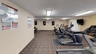 This fitness center offers a comprehensive workout environment spread across a single level. The space is divided into distinct functional zones, each outfitted with a variety of exercise equipment to accommodate different training needs.

The main cardio area features a row of treadmills and elliptical trainers from brands including Matrix, providing ample options for aerobic exercise. Motivational athletic posters and instructional fitness charts are displayed along the neutral-toned walls, adding visual interest while serving as practical references. A wall-mounted television is positioned for easy viewing during workouts.

A separate strength training area houses a multi-station cable machine along with additional resistance equipment, giving users access to a range of resistance-based exercises. Protective rubber mats are placed beneath the equipment throughout the space.

A small seating nook with a bistro-style table and bar stools offers a rest area, accompanied by a wall-mounted air conditioning unit for climate control. Vertical blinds cover the windows, allowing natural light while maintaining privacy. Overhead fluorescent panels provide consistent illumination throughout, and patterned commercial carpet covers the floors, lending the space a practical, durable finish.