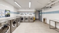 Located at 70 Spadina Rd in Toronto's Annex neighbourhood, this shared laundry facility offers a clean, well-organized space designed for efficient, high-capacity use. The room is lined with a row of modern front-load washing machines and dryers finished in a sleek metallic grey, providing ample capacity for residents. The walls feature a crisp white tile finish accented by a horizontal band of light blue subway tiles that runs the perimeter of the room, lending a clean and cohesive aesthetic. Large-format light grey floor tiles contribute to the bright, hygienic feel of the space. A dedicated utility area includes a white cabinet-base unit topped with a stainless steel sink, accompanied by visible plumbing infrastructure along the walls. A folding table with a chair provides a convenient surface for sorting and folding laundry. Overhead, fluorescent strip lighting ensures the space is well-illuminated throughout. Exposed pipework running along the ceiling reflects the functional, utilitarian nature of the room. A grey access door at the far end provides a secondary entry point. The space is practical and thoughtfully equipped to serve the needs of building residents.
