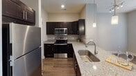 This thoughtfully designed single-floor residence combines modern finishes with a warm, comfortable ambiance. The kitchen is a standout feature, showcasing rich dark cabinetry with sleek hardware, granite countertops, a subway tile backsplash, and a full suite of stainless steel appliances including a refrigerator, range, and microwave. A undermount stainless sink sits beneath elegant pendant lighting, adding a refined touch to the space.

The open living area is bright and welcoming, bathed in natural light from a generous set of large windows fitted with horizontal blinds. Neutral-toned walls and rich, dark hardwood-style flooring flow seamlessly throughout the space, creating a cohesive and inviting atmosphere. The room offers ample space for comfortable living and is complemented by recessed lighting and a track light fixture.

The bathroom is well-finished and functional, featuring granite countertops atop dark cabinetry, a large mirror, and a tub-shower combination enclosed by sliding glass doors. Floor-to-ceiling tile surrounds the tub, while the hardwood-style flooring continues into this space for a consistent aesthetic. Overall, the residence presents a clean, contemporary style with quality finishes at every turn.