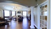Located at 3655 Rosslare Harbour Dr in Pickerington, Ohio, this single-floor space presents a refined and inviting atmosphere defined by thoughtful architectural detailing and warm, classic finishes. Rich, dark hardwood flooring runs throughout, anchoring the space with a sense of elegance and continuity.

The main lounge area is a standout feature, highlighted by a traditional fireplace with a white mantle, above which a mounted television serves as a central focal point. Comfortable seating arrangements are positioned to encourage conversation and relaxation, while large windows along the walls allow ample natural light to fill the room. Two decorative chandeliers and a ceiling fan overhead add a layered, classic lighting aesthetic that enhances the warm ambiance.

A distinctive white decorative column with a detailed capital separates the lounge from an adjoining bar area, which is outfitted with high-top bar seating and a dark wood back bar with extensive shelving for storage and display. Arched transitions and tray ceiling details add architectural character throughout, giving the space a polished, well-appointed feel.

Overall, the space balances classic design elements with practical functionality, creating an environment that is both visually impressive and well-suited for gathering and hospitality.