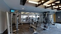 This fitness center offers a comprehensive workout environment designed to accommodate a wide range of exercise needs. The space is defined by its modern aesthetic, featuring deep gray walls that create a focused, energetic atmosphere. Exposed dark wood beam accents along the ceiling add architectural character, while contemporary linear pendant lighting ensures the area is well-lit throughout.

A large floor-to-ceiling mirror spans one wall, visually expanding the space and allowing users to monitor their form during workouts. The layout is thoughtfully organized, housing an impressive selection of strength training equipment including cable machines, weight racks, plate-loaded machines, and adjustable benches. Free weights and barbells are conveniently accessible, catering to both beginner and advanced fitness routines.

The flooring consists of durable, textured carpet tiles suited for a high-traffic fitness environment. Recessed lighting supplements the pendant fixtures, ensuring even illumination throughout. The overall design balances functionality with a polished, contemporary look, making it a practical and inviting space for daily exercise.