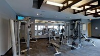 This fitness center offers a comprehensive workout environment designed to accommodate a wide range of exercise needs. The space is defined by its modern aesthetic, featuring deep gray walls that create a focused, energetic atmosphere. Exposed dark wood beam accents along the ceiling add architectural character, while contemporary linear pendant lighting ensures the area is well-lit throughout.

A large floor-to-ceiling mirror spans one wall, visually expanding the space and allowing users to monitor their form during workouts. The layout is thoughtfully organized, housing an impressive selection of strength training equipment including cable machines, weight racks, plate-loaded machines, and adjustable benches. Free weights and barbells are conveniently accessible, catering to both beginner and advanced fitness routines.

The flooring consists of durable, textured carpet tiles suited for a high-traffic fitness environment. Recessed lighting supplements the pendant fixtures, ensuring even illumination throughout. The overall design balances functionality with a polished, contemporary look, making it a practical and inviting space for daily exercise.