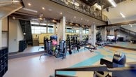 This two-story fitness facility is designed to accommodate a wide range of workout needs within a generously proportioned, open-plan environment. The main floor is anchored by an impressive array of strength training machines, free weights, cable systems, and resistance equipment, all arranged across a distinctive multi-colored vinyl floor featuring bold teal, orange, and yellow geometric patterns. Large structural columns define the layout while full-length mirrored walls visually expand the space and allow users to monitor their form throughout workouts. Multiple flat-screen televisions are mounted throughout the floor for entertainment. Cardio equipment, including stationary and spin cycles, is positioned near the mirrored walls. The upper mezzanine level, accessible via a metal staircase, overlooks the main floor and is framed by an expansive band of floor-to-ceiling windows that flood the space with natural light and offer views of the surrounding landscape. Exposed ductwork and industrial-style ceiling fixtures contribute to a modern, utilitarian aesthetic. The second floor also provides open workout space with additional equipment and wall-mounted artwork, creating a motivating and dynamic fitness environment across both levels.