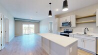 This well-appointed 2-bedroom, 2-bathroom residence in Hastings, MN offers a thoughtfully designed open-concept layout with modern finishes throughout. The kitchen is a standout feature, showcasing white shaker-style cabinetry with matte black hardware, crisp white countertops, a full suite of stainless steel appliances including a range, microwave, dishwasher, and refrigerator, and a generously sized center island with pendant lighting overhead. The kitchen flows seamlessly into the living room, which is bathed in natural light and opens directly onto a private balcony through sliding glass doors, offering outdoor views of the surrounding area. Light wood-tone luxury vinyl plank flooring extends throughout the main living spaces, lending a clean and contemporary feel. Both bedrooms are carpeted and well-proportioned, with the secondary bedroom featuring a window with outdoor views and a dedicated closet. The two bathrooms are finished with gray vanities, white countertops, and wood-tone LVP flooring, with one featuring a tub-shower combination and the other offering an extended vanity with ample cabinetry. A connecting hallway ties the layout together efficiently, and two closets provide practical storage solutions throughout the residence.
