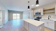 This well-appointed 2-bedroom, 2-bathroom residence in Hastings, MN offers a thoughtfully designed open-concept layout with modern finishes throughout. The kitchen is a standout feature, showcasing white shaker-style cabinetry with matte black hardware, crisp white countertops, a full suite of stainless steel appliances including a range, microwave, dishwasher, and refrigerator, and a generously sized center island with pendant lighting overhead. The kitchen flows seamlessly into the living room, which is bathed in natural light and opens directly onto a private balcony through sliding glass doors, offering outdoor views of the surrounding area. Light wood-tone luxury vinyl plank flooring extends throughout the main living spaces, lending a clean and contemporary feel. Both bedrooms are carpeted and well-proportioned, with the secondary bedroom featuring a window with outdoor views and a dedicated closet. The two bathrooms are finished with gray vanities, white countertops, and wood-tone LVP flooring, with one featuring a tub-shower combination and the other offering an extended vanity with ample cabinetry. A connecting hallway ties the layout together efficiently, and two closets provide practical storage solutions throughout the residence.