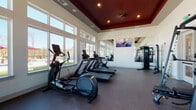 Located at 575 Concord Rd in Jefferson, GA, this fitness center offers a thoughtfully designed single-floor workout facility that balances functionality with an inviting atmosphere. The space is defined by its striking tray ceiling finished in a rich, dark red tone and accented with recessed lighting and a ceiling fan, creating a polished yet energetic ambiance throughout.

Large, floor-to-ceiling windows line the walls, flooding the interior with abundant natural light and offering direct views of the outdoor pool area and surrounding pergola, making workouts feel open and refreshing. Full-length mirrors are strategically placed to amplify the sense of space and allow users to monitor their form during exercise.

The cardio zone is comprehensively outfitted with treadmills, ellipticals, stationary bikes, and rowing machines — including Precor equipment — all positioned to take advantage of the exterior views. A flat-screen television is wall-mounted for entertainment during sessions.

The strength training area features a multi-functional cable machine, a Smith machine, adjustable benches, and a well-stocked dumbbell rack, catering to a wide range of resistance training needs. Durable rubber flooring covers the entire space, providing comfort and safety underfoot. The fitness center is accessed through glass-panel doors, reinforcing its bright, transparent aesthetic and seamlessly connecting it to the broader community amenity area.