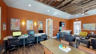 This single-floor amenity space blends functional design with striking aesthetic character. The interior is immediately distinguished by its vaulted ceiling adorned with reclaimed wood planks, which contrast warmly against crisp white trim and create a rustic yet polished ambiance throughout the room.

Bold terracotta-orange walls serve as the defining design element, lending the space an energetic and welcoming atmosphere. The flooring consists of rich, dark hardwood planks that ground the space with a sense of warmth and durability.

A dedicated business center area features a built-in workstation with multiple desktop computers and ergonomic seating, making it well-suited for productivity. Decorative wall sconces and colorful abstract artwork adorn the walls, adding a curated, gallery-like quality to the environment.

A kitchenette area is integrated along one wall, finished with teal-gray cabinetry, a compact sink, and countertop appliances, providing convenient refreshment options. A wall-mounted flat-screen television is positioned nearby for media use.

A collaborative table with seating occupies the central area, offering a versatile space for meetings or group work. A glazed interior door provides a sense of openness while maintaining separation between zones. Recessed lighting supplements the sconces, ensuring the space is well-lit throughout.