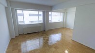 This single-floor space offers a spacious, open layout characterized by its highly polished, checkerboard-patterned parquet flooring that reflects natural light throughout the room. Two large windows dressed with horizontal blinds dominate the exterior wall, flooding the interior with abundant daylight while providing views of the surrounding urban streetscape. The walls are finished in clean white, contributing to a bright and airy atmosphere that enhances the sense of openness. Baseboard radiator units are positioned beneath the windows, indicating a functional heating system integrated into the space. The layout is open and unobstructed, offering considerable flexibility for a variety of uses or configurations. A doorway visible along the far wall suggests additional access points or adjoining spaces beyond the main room. The overall aesthetic is straightforward and utilitarian, with neutral finishes and minimal ornamentation, making it a versatile canvas for a range of purposes. The combination of natural light, generous floor space, and polished flooring gives the space a clean, functional character well-suited to customization.