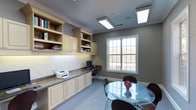 This single-floor office space is thoughtfully designed to support both focused work and small group collaboration. The room is anchored by a long built-in workstation with a granite-style countertop, flanked by cream-colored cabinetry and open shelving above — providing ample storage for books, binders, and office essentials. The workspace accommodates a desktop computer and printer, keeping daily operations organized and efficient.

Natural light filters in through a large multi-pane window, offering a pleasant view of the surrounding landscape and contributing to a bright, comfortable atmosphere. The space is further illuminated by recessed ceiling light panels that ensure consistent, even lighting throughout the room.

A circular glass-top dining or conference table with sleek dark chairs sits opposite the workstation, creating a dedicated area for meetings or collaborative discussions. Soft carpeting underfoot adds warmth and acoustic comfort to the overall environment.

The walls are finished in a cool gray tone that complements the cream cabinetry, while crown molding along the ceiling adds a refined architectural detail. This space strikes a balance between professional functionality and comfortable design, making it a versatile and well-equipped work environment.