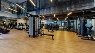 Located at 375 Camino De La Reina in San Diego's Mission Valley, this contemporary fitness center is designed to deliver a premium workout experience across a spacious, open floor plan. The facility is anchored by soaring double-height ceilings in the main training area, with floor-to-ceiling glass walls and large windows that flood the space with natural light and provide views of the surrounding exterior. Warm hardwood-style flooring runs throughout, complementing the sleek, industrial-modern aesthetic defined by exposed concrete ceilings, white subway tile accent walls, and linear pendant lighting.

The main floor accommodates a comprehensive array of cardio equipment, including treadmills and cycling machines, alongside multi-station cable machines, weight benches, and a full dumbbell rack. A dedicated functional training zone features TKO heavy punching bags, battle ropes, TRX suspension trainers, medicine balls, and stability equipment, offering diverse training modalities. A glass-partitioned secondary area houses additional strength machines, including leg press and seated resistance equipment, and is equipped with a dual water fountain station for convenience. Wall-to-ceiling mirrors throughout amplify the sense of space and support proper form during workouts. The overall design balances high-end aesthetics with serious athletic functionality.