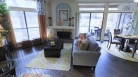 This single-story living space offers a well-appointed and thoughtfully designed interior centered around a striking fireplace framed by a decorative arched alcove painted in a soft blue hue, which serves as a natural focal point for the room. The fireplace surround features a textured stone facade and a clean white mantel, adding both warmth and architectural character. Rich, dark hardwood flooring runs throughout, grounding the space and providing a warm contrast to the lighter wall tones. Large windows along the perimeter flood the room with generous natural light, while offering views of an outdoor patio area with additional seating visible beyond. The living area is furnished with a spacious gray sectional sofa accented by colorful coral and green throw pillows, complemented by a bold geometric yellow-and-white area rug that adds a lively, contemporary energy. A substantial dark wood coffee table anchors the seating arrangement. Drapery in warm orange and patterned white tones frames the windows, adding texture and visual interest. The overall ambiance is inviting and vibrant, blending eclectic décor elements with comfortable, functional furnishings in a bright, open setting.