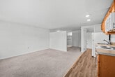 This single-floor residential unit offers a well-organized layout with distinct living, sleeping, kitchen, and bathroom areas. The kitchen is equipped with warm-toned wooden cabinetry, white appliances including a refrigerator, electric range, and built-in microwave, complemented by a classic subway tile backsplash and a double-basin sink. A door in the kitchen area provides convenient exterior access and allows natural light to filter in through adjacent windows. The living area features neutral, plush carpeting and a partial wall divider that subtly separates spaces while maintaining an open feel. A peek into the bathroom reveals a vanity with lighted mirror and cabinetry, adding a functional touch. The bedroom is generously proportioned, offering soft carpet underfoot and a large window fitted with horizontal blinds that invite natural light and outdoor views. Baseboard heating runs throughout the carpeted rooms, ensuring year-round comfort. A bedroom closet with sliding panel doors provides ample storage space, with additional shelving visible within. Wall sconce lighting adds a warm ambiance to the sleeping area. Overall, the unit presents a clean, practical, and comfortable living environment with consistent neutral tones and straightforward, functional finishes throughout.