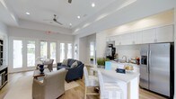 Located at 2440 Salinger Ct in Wilmington, North Carolina, this single-floor community space offers a welcoming and well-appointed environment designed for both comfort and functionality. The open-concept layout seamlessly connects a relaxed lounge area with a fully equipped kitchenette, creating an inviting atmosphere for gathering and socializing. The lounge is furnished with a navy blue sofa and wicker accent chairs arranged around a central coffee table, providing a comfortable and stylish seating arrangement. Large French doors along the exterior wall flood the space with generous natural light and offer views of the surrounding greenery, enhancing the bright, airy feel throughout. The kitchenette features crisp white cabinetry, a light-toned countertop with bar seating, and modern appliances, making it a practical and attractive amenity. Recessed lighting and a ceiling fan complement the tray ceiling, adding architectural interest while maintaining a clean, contemporary aesthetic. Light hardwood-style flooring runs throughout, tying the space together with warmth and continuity. Overall, the space strikes a balanced tone between casual comfort and polished design, making it an ideal communal hub.