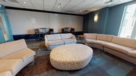 This single-floor lounge space offers a well-appointed and contemporary environment designed for comfort and social interaction. The room is defined by its teal and neutral color palette, with deep teal accent walls contrasting against lighter beige and off-white surfaces, creating a modern yet inviting atmosphere.

The seating area serves as the focal point of the space, featuring multiple curved and rounded sectional sofas upholstered in soft neutral tones, arranged around a large oval ottoman with a distinctive abstract patterned fabric. This central grouping is oriented toward a large wall-mounted flat-screen television, supported by a sleek, low-profile media console with integrated storage. Built-in acoustic panels flank the TV wall, enhancing both the aesthetics and sound quality of the room.

Natural light filters in through windows positioned along one wall, offering exterior views and brightening the space. Wall sconces with frosted glass shades provide warm supplemental lighting throughout. A drop ceiling with recessed lighting adds to the clean, finished look overhead.

Along the opposite side of the room, high-top tables paired with dark bar-height chairs offer an alternative seating arrangement, ideal for casual meetings or dining. The floor is covered in multi-toned modular carpet tiles in shades of teal, brown, and gray, adding visual texture and depth to the overall design.