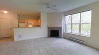 This single-floor space offers a thoughtfully designed open layout that seamlessly connects the living area and kitchen. The living room is a comfortable, carpeted space anchored by a tile-surround fireplace, which serves as a focal point along the main wall. A ceiling fan overhead provides both practical airflow and a subtle design element, while a large window along one wall allows generous natural light to filter in, offering pleasant views of surrounding greenery. A baseboard heating unit beneath the window ensures year-round comfort.

The kitchen is accessible via an open pass-through counter, creating a connected feel between the cooking and living spaces. Warm wood-toned cabinetry lines the kitchen walls, complemented by track lighting mounted on the ceiling above, which illuminates the workspace effectively. A built-in microwave and light-colored countertops complete the functional kitchen setup. The pass-through bar counter also provides casual seating or serving opportunities, enhancing the open, social nature of the layout. Overall, the space presents a bright, inviting atmosphere with a clean, neutral palette throughout.