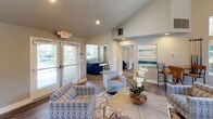 Located at 7952 Pocket Rd in Sacramento, California, this single-story community clubhouse offers a well-appointed gathering space designed for both comfort and functionality. The entry lounge welcomes visitors with vaulted ceilings, recessed lighting, and wood-style flooring throughout, creating a bright and open atmosphere. Comfortable patterned seating arrangements and tasteful coastal-inspired artwork set a relaxed, inviting tone.

A large communal dining and gathering area features an expansive natural wood bar-height table with modern chrome seating, positioned adjacent to a fully equipped kitchen. The kitchen is outfitted with dark cabinetry, stainless steel appliances including a gas range and French door refrigerator, a stone tile backsplash, and quartz countertops — ideal for hosting events and gatherings.

The clubhouse also includes a dedicated media lounge, where sliding glass barn doors partition a cozy space furnished with a plush blue sofa and large wall-mounted flat-screen TV. A connected sitting area features additional seating and colorful accent artwork. A sunlit nook lined with expansive windows offers views of the surrounding landscaped grounds and pool area, providing an ideal spot for quiet work or relaxation. Together, these spaces form a versatile, well-designed community hub.