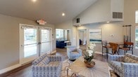 Located at 7952 Pocket Rd in Sacramento, California, this single-story community clubhouse offers a well-appointed gathering space designed for both comfort and functionality. The entry lounge welcomes visitors with vaulted ceilings, recessed lighting, and wood-style flooring throughout, creating a bright and open atmosphere. Comfortable patterned seating arrangements and tasteful coastal-inspired artwork set a relaxed, inviting tone.

A large communal dining and gathering area features an expansive natural wood bar-height table with modern chrome seating, positioned adjacent to a fully equipped kitchen. The kitchen is outfitted with dark cabinetry, stainless steel appliances including a gas range and French door refrigerator, a stone tile backsplash, and quartz countertops — ideal for hosting events and gatherings.

The clubhouse also includes a dedicated media lounge, where sliding glass barn doors partition a cozy space furnished with a plush blue sofa and large wall-mounted flat-screen TV. A connected sitting area features additional seating and colorful accent artwork. A sunlit nook lined with expansive windows offers views of the surrounding landscaped grounds and pool area, providing an ideal spot for quiet work or relaxation. Together, these spaces form a versatile, well-designed community hub.