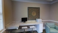 This single-floor space offers a thoughtfully designed interior that blends practicality with refined aesthetics. The standout feature is a built-in workstation finished with light granite countertops, providing ample surface area for productivity. The countertop is supported by crisp white cabinetry below, offering generous storage while maintaining a clean, polished look. Two modern chrome-legged bar stools are tucked neatly beneath the counter, creating a comfortable and versatile workspace. The walls are finished in a warm, neutral taupe tone that lends a calm and welcoming ambiance throughout the space. Crown molding lines the ceiling, adding a classic architectural detail that elevates the overall design. A striking large-format decorative wall panel with an intricate mandala-style motif in deep tones and gold accents serves as the focal point above the workstation, infusing the space with artistic character. The flooring features a rich dark surface that grounds the room and complements the lighter countertops and wall tones. Overall, this space effectively combines a professional, functional layout with tasteful design elements, making it suitable for both work and welcoming guests.