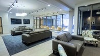 This single-floor space is designed as a contemporary communal lounge, thoughtfully arranged to accommodate both relaxed socializing and casual work or dining. The interior is anchored by a seating area furnished with dark leather sofas and armchairs, arranged around a low coffee table atop a area rug over light hardwood-style flooring. Track lighting runs across the ceiling, supplemented by recessed fixtures and pendant lights, ensuring the space is well-lit throughout.

Toward the rear, floor-to-ceiling glass windows and doors flood the space with abundant natural light, creating an open, airy atmosphere. This glazed section transitions into a separate dining or collaborative zone, outfitted with round tables and bright yellow chairs — a vibrant contrast to the neutral tones of the lounge area. Glass-paneled partitions help define this zone while maintaining visual connectivity throughout the space.

The overall design blends functional furnishings with a clean, modern aesthetic. Light walls, structured columns, and the interplay of natural and artificial light contribute to a welcoming, versatile environment suited for a variety of activities.