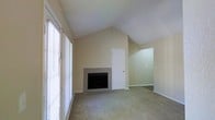 This single-floor residence offers a thoughtfully designed layout that balances openness with comfort. The living area immediately impresses with its dramatic vaulted ceilings that rise to a sharp peak, lending an expansive, airy quality to the space. Neutral beige walls and soft wall-to-wall carpeting create a warm, understated backdrop throughout. A tiled fireplace serves as the focal point of the main living area, framed by clean lines and a simple surround that adds a touch of classic character. Large windows fitted with horizontal blinds line one side of the room, inviting generous natural light while maintaining privacy. An open archway connects the main living space to an adjacent area, enhancing the sense of flow and continuity throughout the floor plan. A white interior door provides access to additional spaces within the home. The overall ambiance is calm and neutral, offering a blank canvas well-suited for a variety of furnishing styles. The combination of elevated ceilings, natural light, and cohesive design elements makes this space feel both functional and inviting.