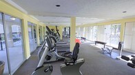This single-floor fitness center offers a comprehensive workout environment designed to accommodate a range of exercise needs. The space is outfitted with an array of cardio equipment, including elliptical trainers, treadmills, and a recumbent stationary bike, all arranged to allow for comfortable circulation throughout the room. Strength training equipment and free weights are also present, providing options for a full-body workout routine. Large floor-to-ceiling windows line the perimeter of the space, flooding the interior with natural light and offering views of the surrounding outdoor environment. The bright, open atmosphere is further enhanced by the soft yellow-toned walls and a textured ceiling, contributing to an airy and welcoming ambiance. A structural column centrally positioned in the room adds an architectural element without impeding the overall flow of the space. Mirrored walls on one side visually expand the room while allowing users to monitor their form during exercise. The carpeted flooring provides comfort underfoot, while exercise mats are available for stretching and floor-based workouts. Overall, this fitness center is thoughtfully arranged to support a productive and comfortable exercise experience.