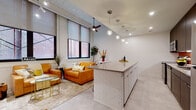 Located at 1144 E Market St in Akron, Ohio, this single-floor unit showcases a thoughtfully designed open-concept layout that blends modern finishes with industrial-inspired character. Exposed white-painted brick walls and structural beam details lend an urban loft aesthetic throughout the space, while light concrete-style flooring ties each area together seamlessly.

The living area is anchored by large black-framed windows that frame views of the surrounding historic brick architecture, flooding the space with natural light. A ceiling fan and globe pendant lighting add both function and visual interest to the room.

The kitchen is a standout feature, offering a galley-style cooking zone with dark wood cabinetry, stainless steel appliances, and a vibrant orange subway tile backsplash that adds a bold pop of color. A white brick-faced island with a quartz countertop provides bar seating and additional prep space, with a row of globe pendant lights suspended above.

The bedroom is a calm, cozy retreat featuring neutral tones, warm gold accent lighting, and a large mirror installation above the bed that enhances the sense of space. A ceiling fan adds comfort overhead.

The bathroom is clean and contemporary, featuring light wood-toned cabinetry, a quartz vanity surface, a glass-enclosed walk-in shower with tile surround, and light hardwood-style flooring — completing the space with a refined, spa-like finish.