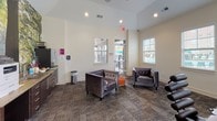 Located at 815 Eden Ave in Fort Mill, South Carolina, this single-story community space is part of the Evolve at Tega Cay property. The open-concept layout is designed to accommodate both collaborative work and relaxed social gatherings, combining functional amenities with a welcoming atmosphere.

The main lounge area features plush, tufted gray armchairs arranged around a sleek glass coffee table, set atop patterned carpet flooring. Multiple windows dressed with white horizontal blinds allow generous natural light to fill the space, creating a bright and airy environment. A glass-panel door provides direct access to an outdoor area, enhancing connectivity to the surrounding community.

A dedicated workstation area is outfitted with a row of desktop computers along a countertop, complemented by a striking full-wall nature mural depicting a lush forest scene. This eye-catching focal point adds depth and visual interest to the room. Vaulted ceilings in this section contribute to an elevated sense of space.

A conference-style table surrounded by dark, modern chairs serves as a central gathering hub, suitable for meetings or group activities. A small kitchenette with dark cabinetry is also present. Throughout the space, neutral walls, recessed lighting, and cohesive furnishings create a polished, community-oriented ambiance.